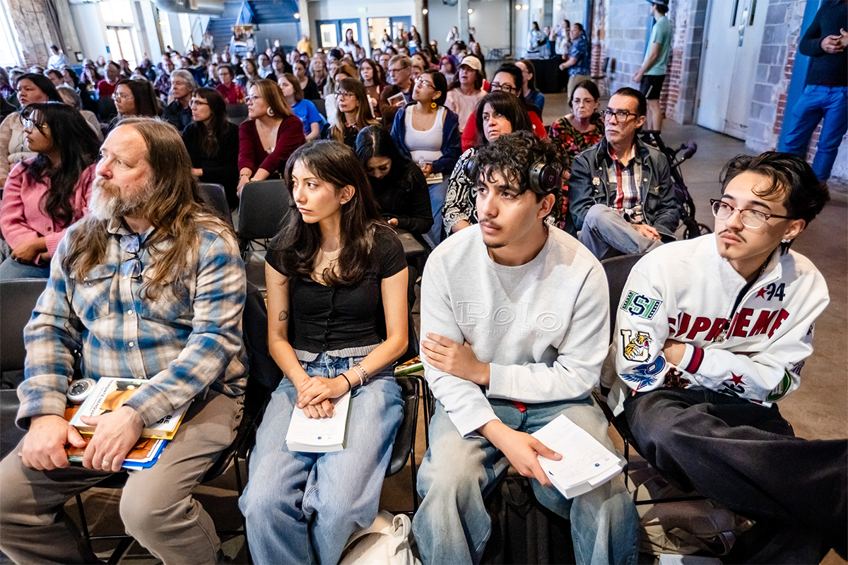 A large audience sits closely together in a hall, listening attentively, with several people in the front row holding books or notebooks.