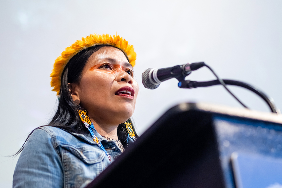 Speaker wearing a yellow headband and traditional earrings speaks into a microphone at a podium.