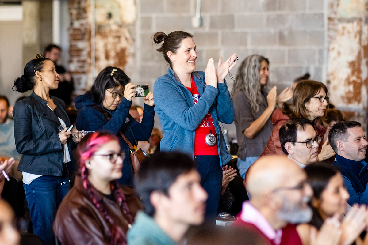 Audience members stand and applaud during an event, with one person in the center clapping while others around them watch and take photos.