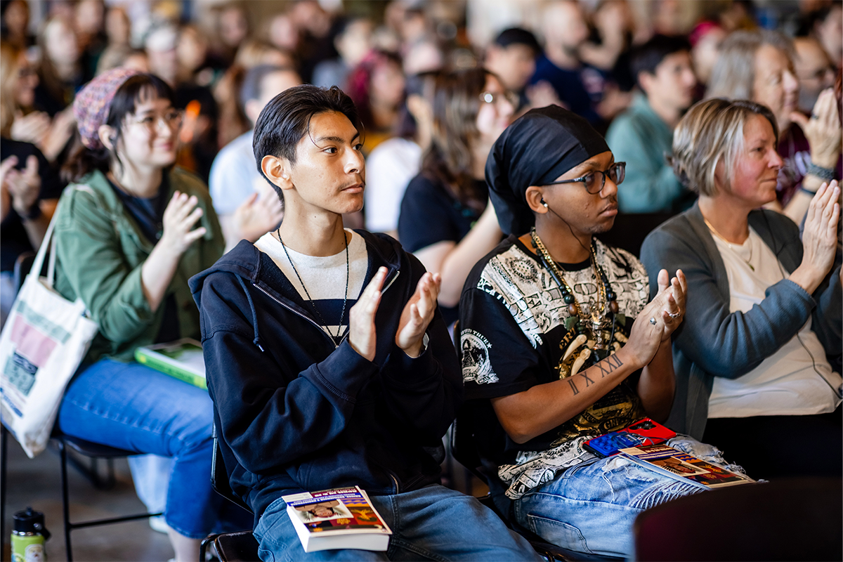 Audience members seated in a crowded event space clap while listening, some holding books in their laps.