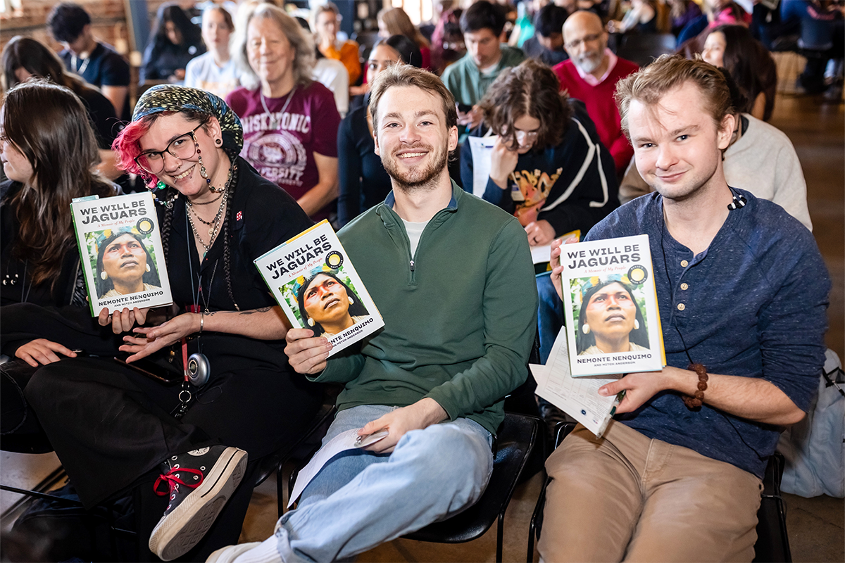 Three attendees sit in a crowded room smiling at the camera while holding copies of a book, with other audience members seated behind them.