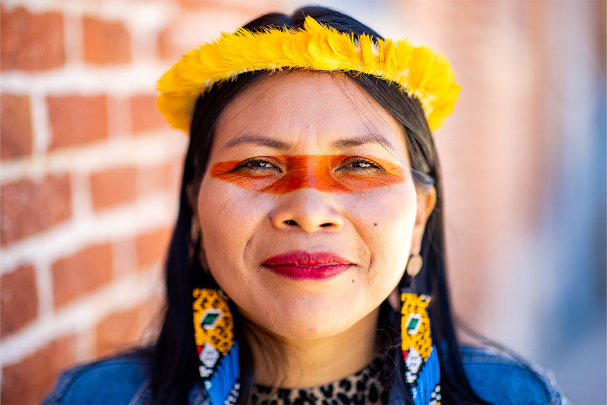 Portrait of a woman wearing a yellow headband, beaded earrings, and red face paint across her eyes, standing in front of a brick wall.