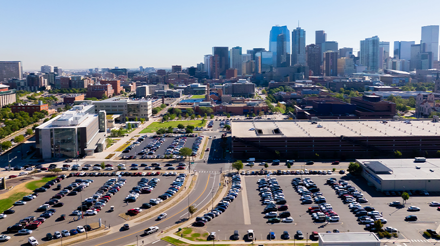 Aerial view of a large parking lot filled with cars near a university campus, with the Denver skyline visible in the background.