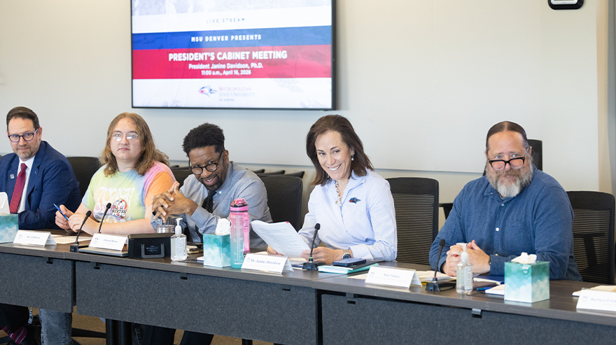 A group of administrators sit at a conference table during a meeting, reviewing documents with a presentation screen behind them displaying “President’s Cabinet Meeting.”