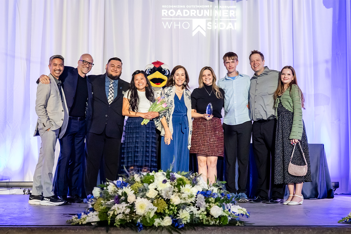 Group of people pose together on stage at an awards event, standing in front of a “Roadrunners Who Soar” backdrop with a university mascot.