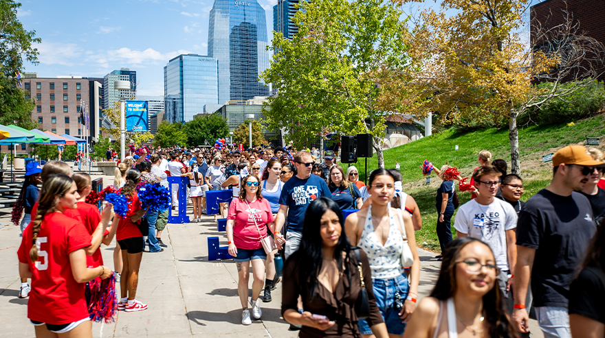Large group of students and attendees walking along a campus pathway during a daytime outdoor event, with cheerleaders and city buildings visible in the background.