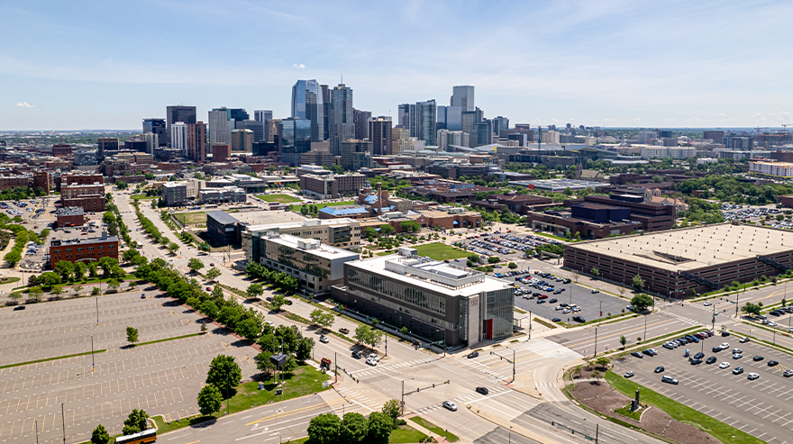 Aerial view of downtown Denver with campus buildings, streets, and parking lots in the foreground under a clear sky.