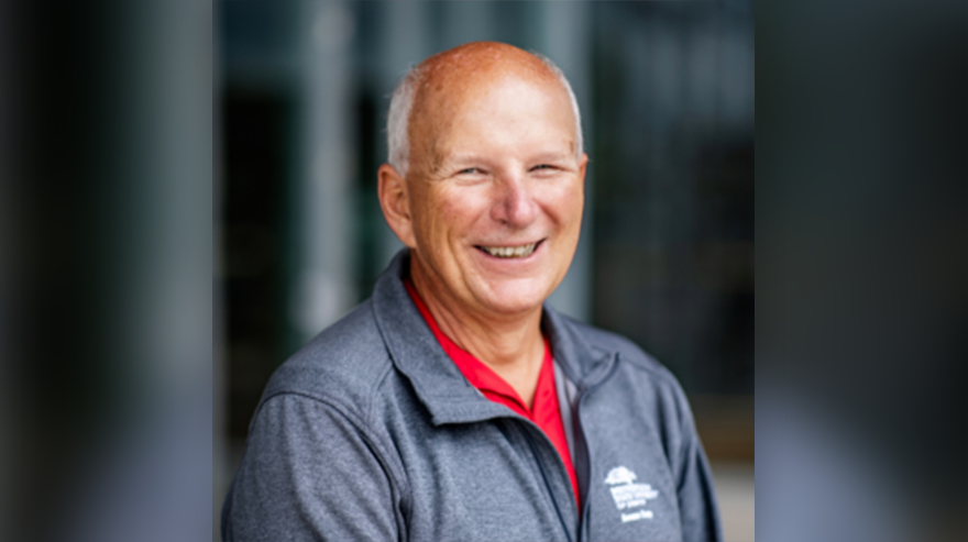 Smiling man in a gray jacket and red shirt posing for a professional portrait, likely associated with an Access Center Director retirement announcement.