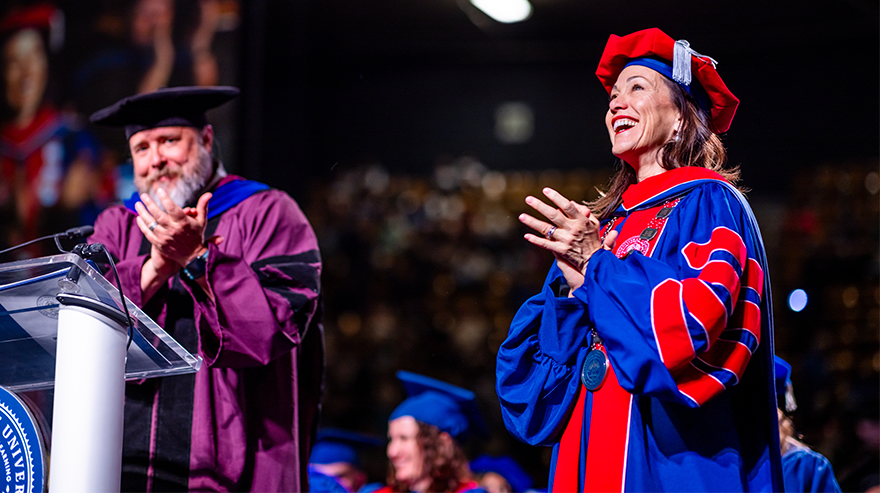 Two faculty members in academic regalia standing on stage and applauding during a commencement ceremony.