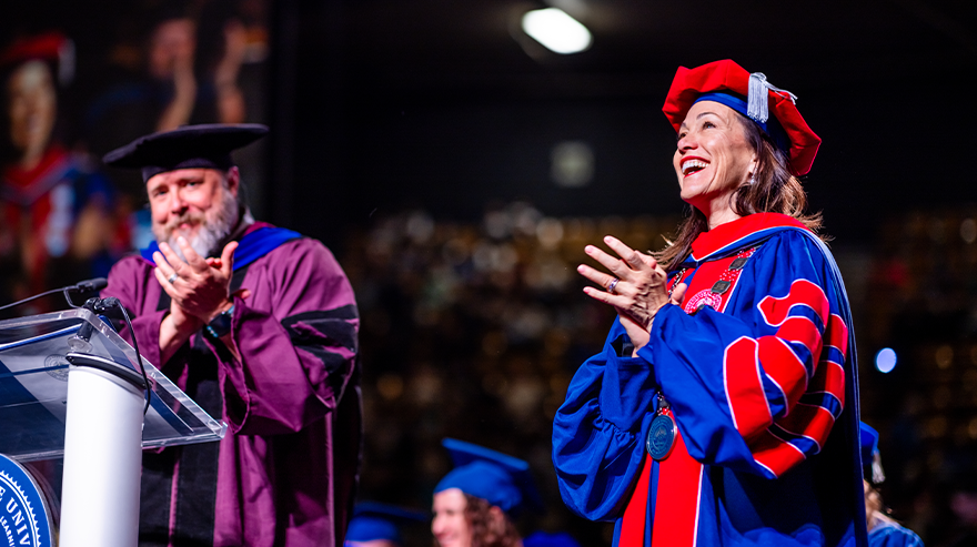 Two faculty members in academic regalia standing on stage and applauding during a commencement ceremony.