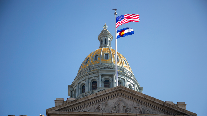The Colorado State Capitol dome with American and Colorado state flags flying against a clear blue sky.