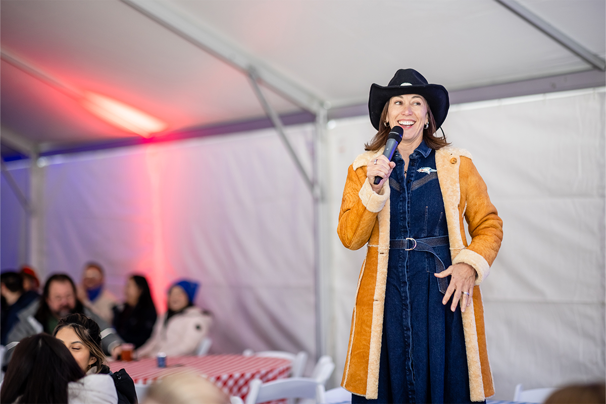 Woman wearing a cowboy hat and western-style outfit speaking into a microphone during an outdoor spring BBQ event under a tent.