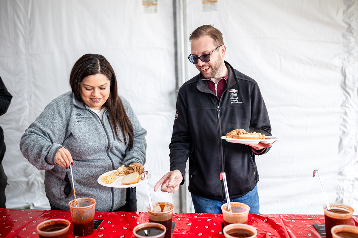Two MSU Denver employees preparing barbecue plates and adding sauce during the Spring 2026 Howdy Rowdy BBQ event at the JSSB Lawn.