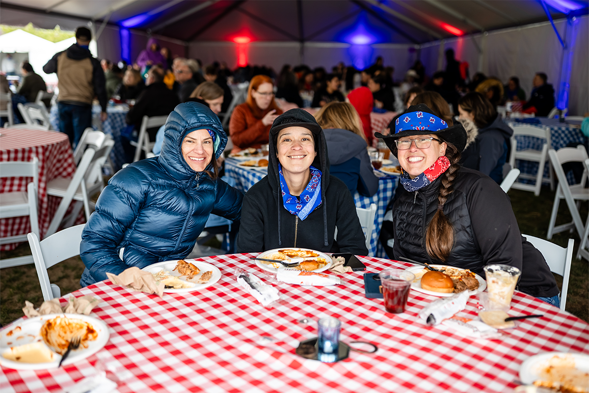 Three MSU Denver community members smiling at a picnic table inside a tent during the Spring 2026 Howdy Rowdy BBQ event at the JSSB Lawn.