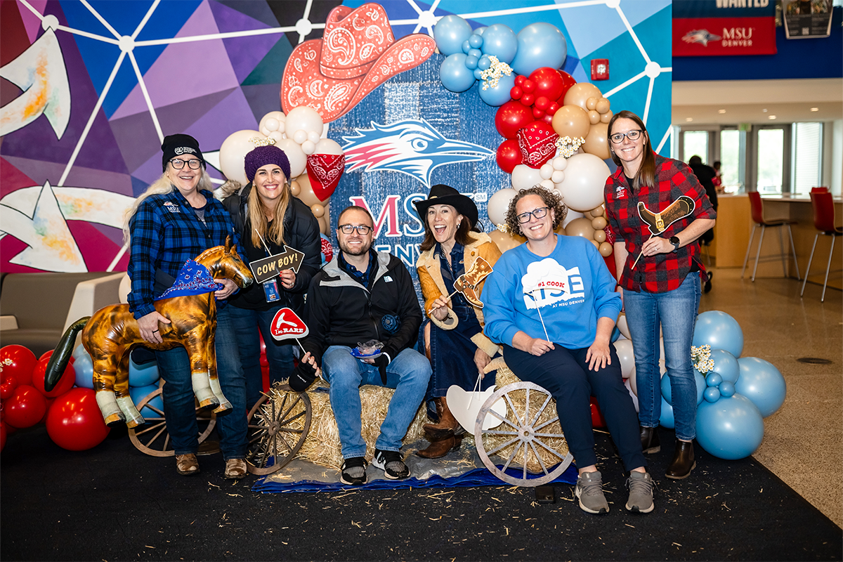 A group of MSU Denver faculty and staff posing with western-themed props in front of a decorated photo backdrop during the Spring 2026 Howdy Rowdy BBQ event.
