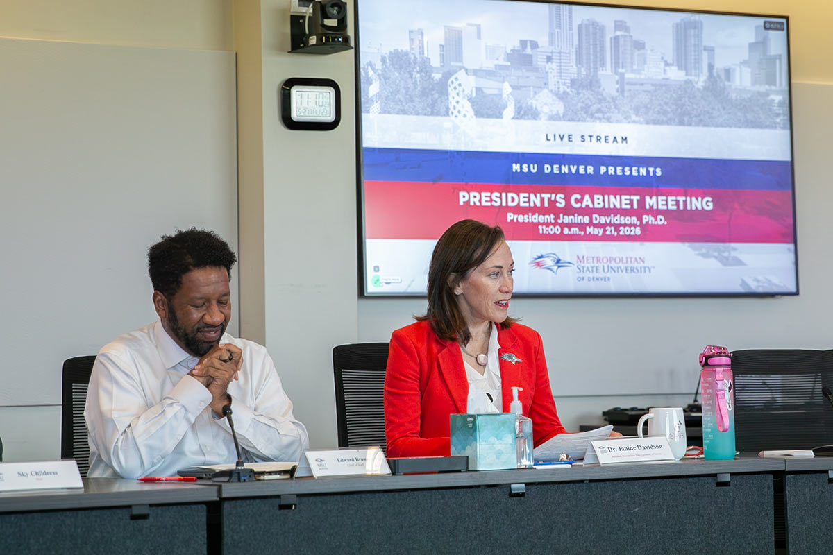 President Janine Davidson and Chief of Staff Edward Brown sit at a meeting table in front of a screen that says "President's Cabinet Meeting"