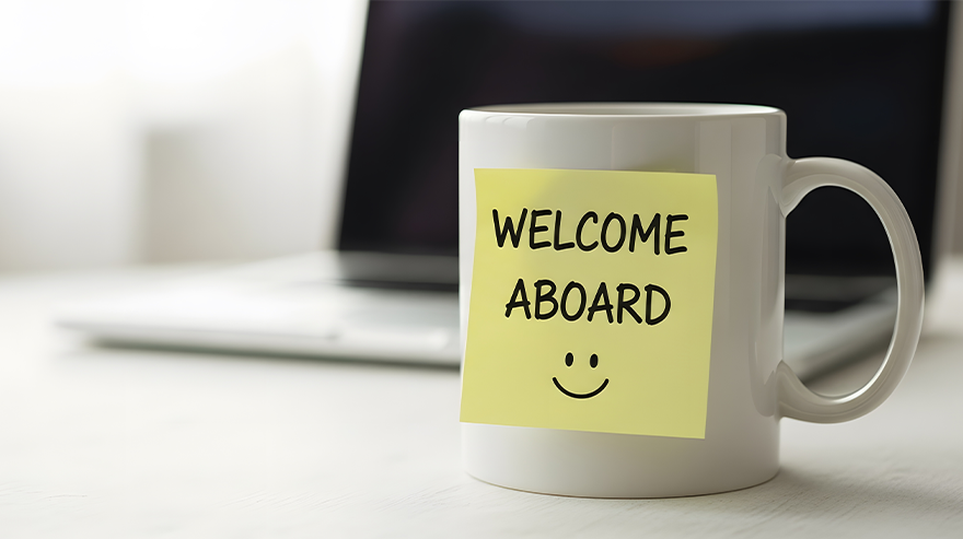 A white mug sitting on a desk with a sticky note over it saying "Welcome Aboard."