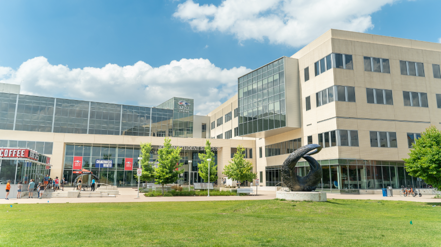 Exterior view of the Jordan Student Success Building on the Auraria Campus, with a grassy lawn, pedestrians, and the campus sculpture visible in front of the building.