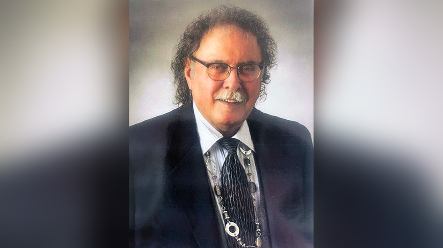 Portrait of retired professor emeritus Charles Angeletti wearing a suit, tie, and glasses, smiling toward the camera.