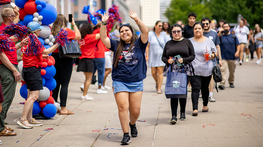 A student wearing blue and MSU Denver-branded clothes with her hands in the air leads a line of other people past a group of University leaders wearing matching red shirts and shaking blue and red pom-poms.