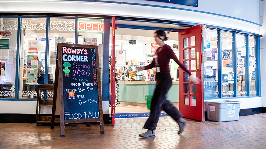 A person walks past the open door of Rowdy's Corner, as well as a sign that says "Rowdy's Corner" and "Food for all."