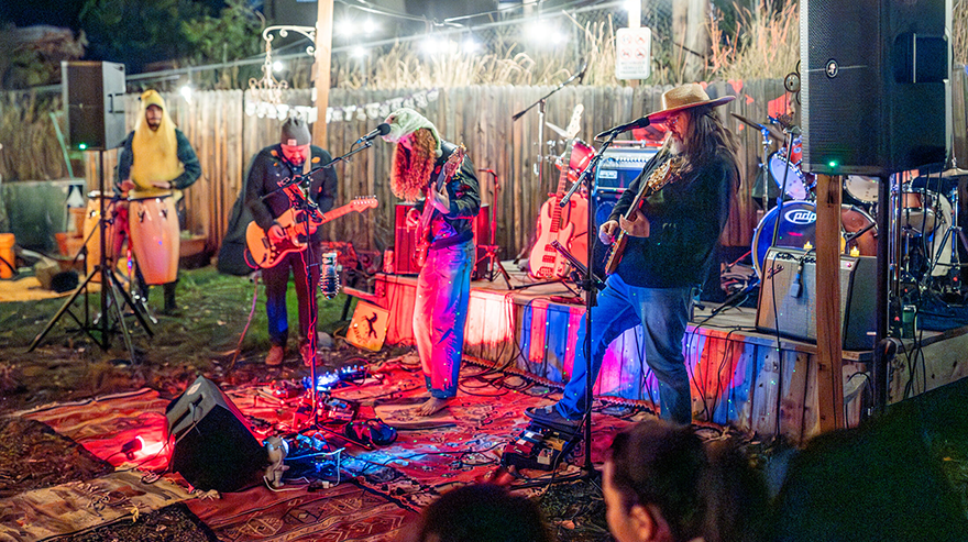 Five musicians play a nighttime show in colorful outfits and costumes under lights in a backyard setting.