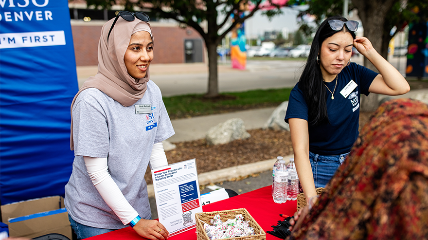 Two MSU Denver students at an outdoor table share promotional materials with branding for the First Generation Initiatives organization in the University Classroom to Career Hub. Photo by Alyson McClaran