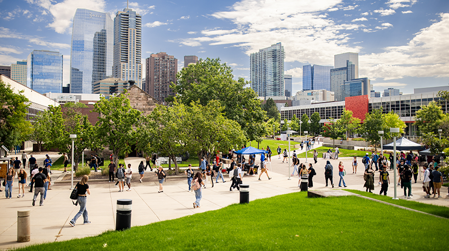 Wide shot of a sidewalk intersection on campus shows students walking amid booths under a blue sky and the Denver skyline in the background