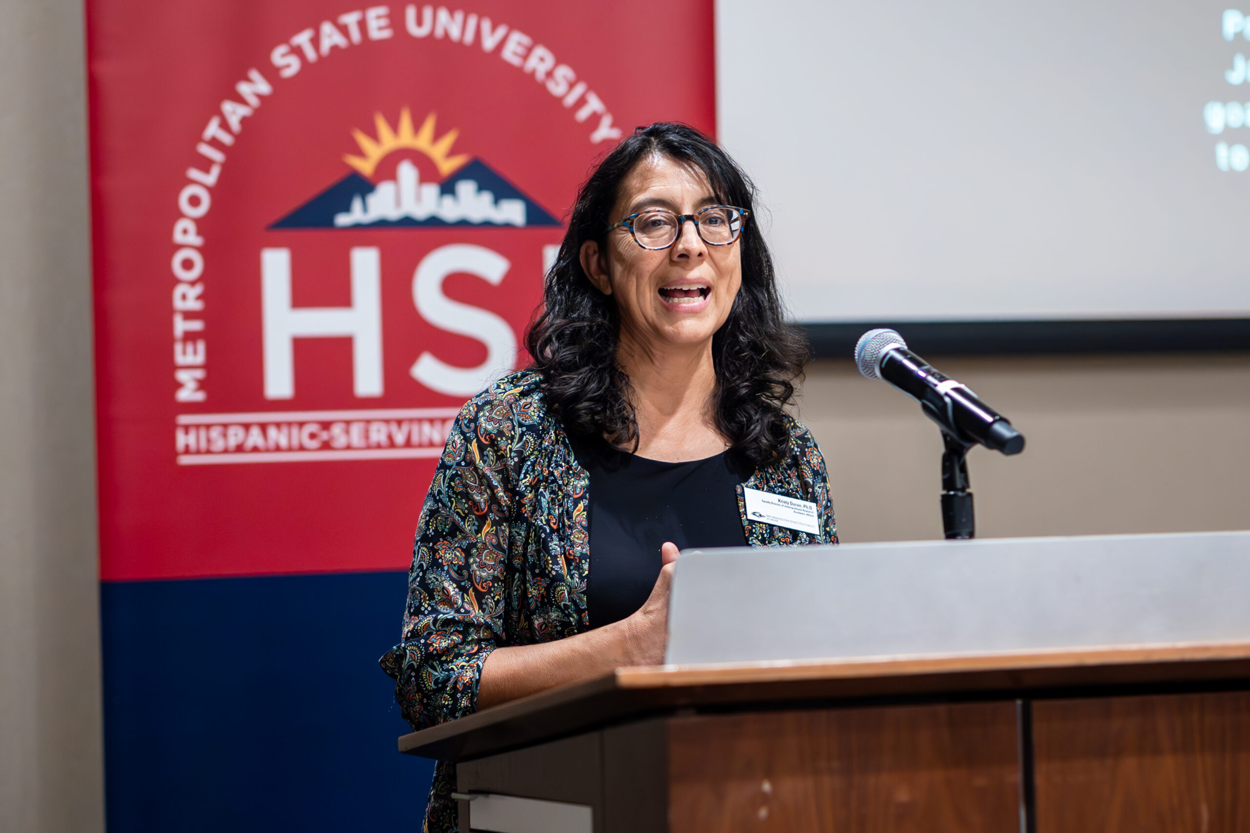 Kristy Duran speaks at a lectern in front of a banner with the MSU Denver Hispanic-Serving Institution logo