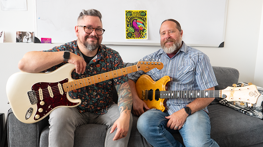 Shawn Meek and Matt Makley pose seated in an office with electric guitars