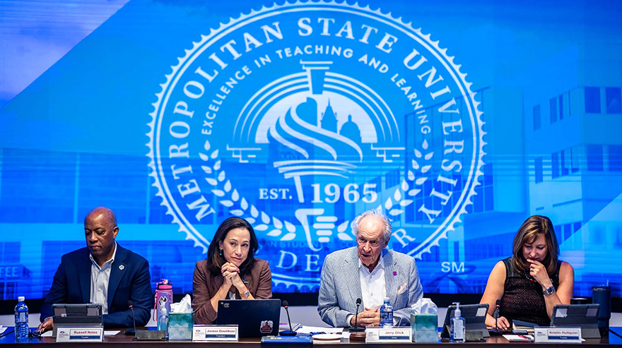 Four University leaders are pictured in front of the MSU Denver crest at the Board of Trustees meeting