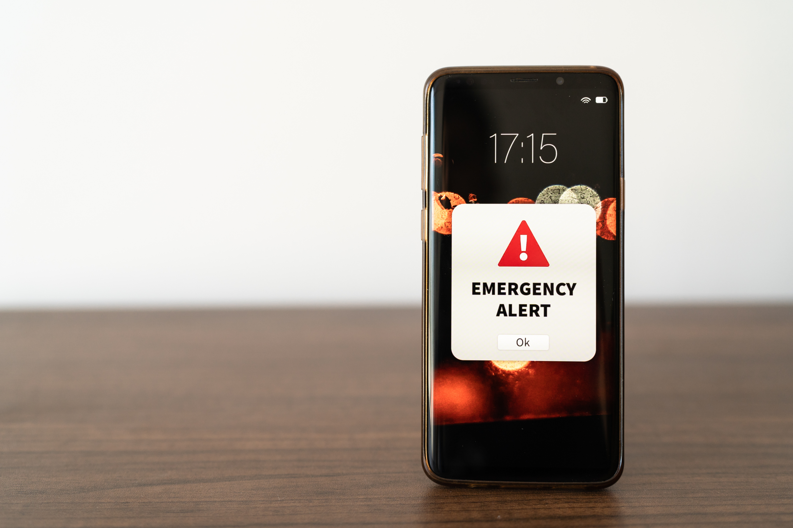 Stock image shows a smartphone standing on a table while showing the message "emergency alert" on the screen.