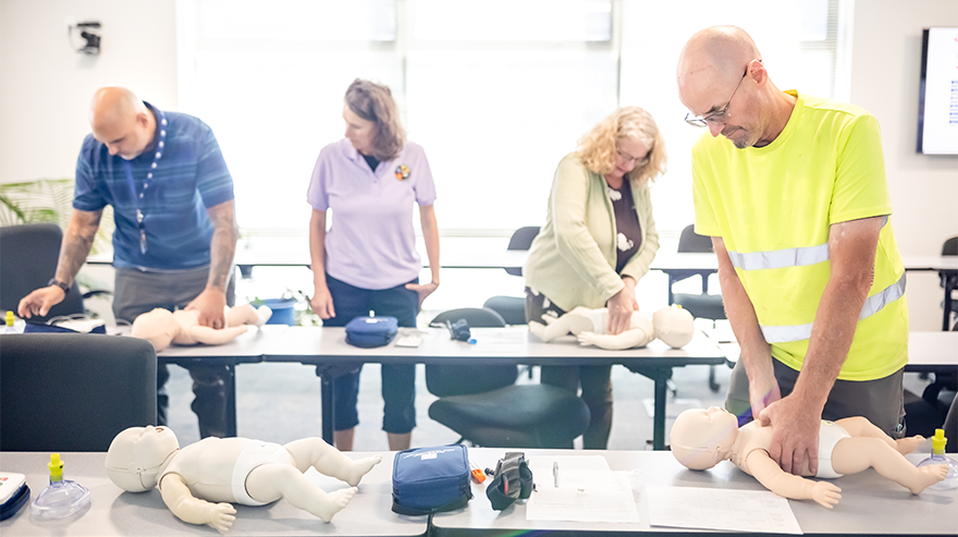 Four people huddle over mannequins for practicing CPR techniques in a well-lit room with tables holding similar equipment