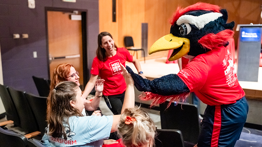 MSU Denver mascot Rowdy high-fives one employee in a group of four who are shown smiling and holding hands up in seats in an auditorium