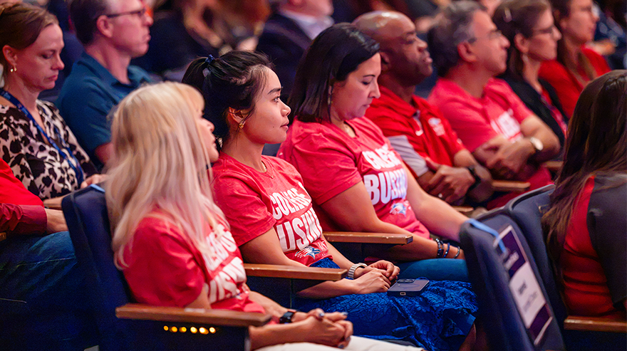 A medium shot shows five people wearing matching College of Business T-shirts in a row in the King Center audience.
