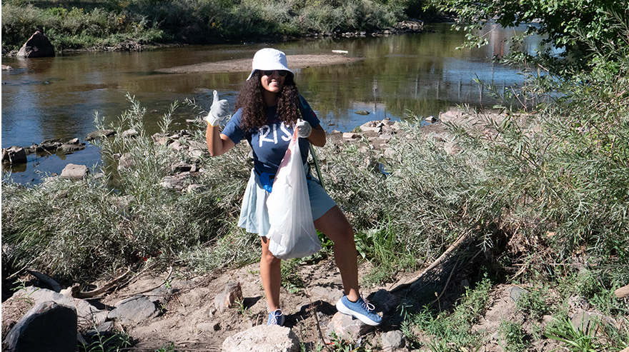 A woman gives a thumbs-up sign while holding a trash bag next to a creek.