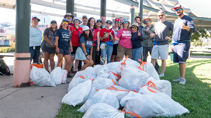 Nineteen people stand around a heaping pile of full trash bags under a park awning.