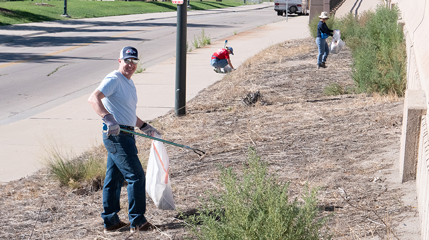 Three people pick up trash with garbage bags and tongs on a strip of land next to a highway and neighborhood road.