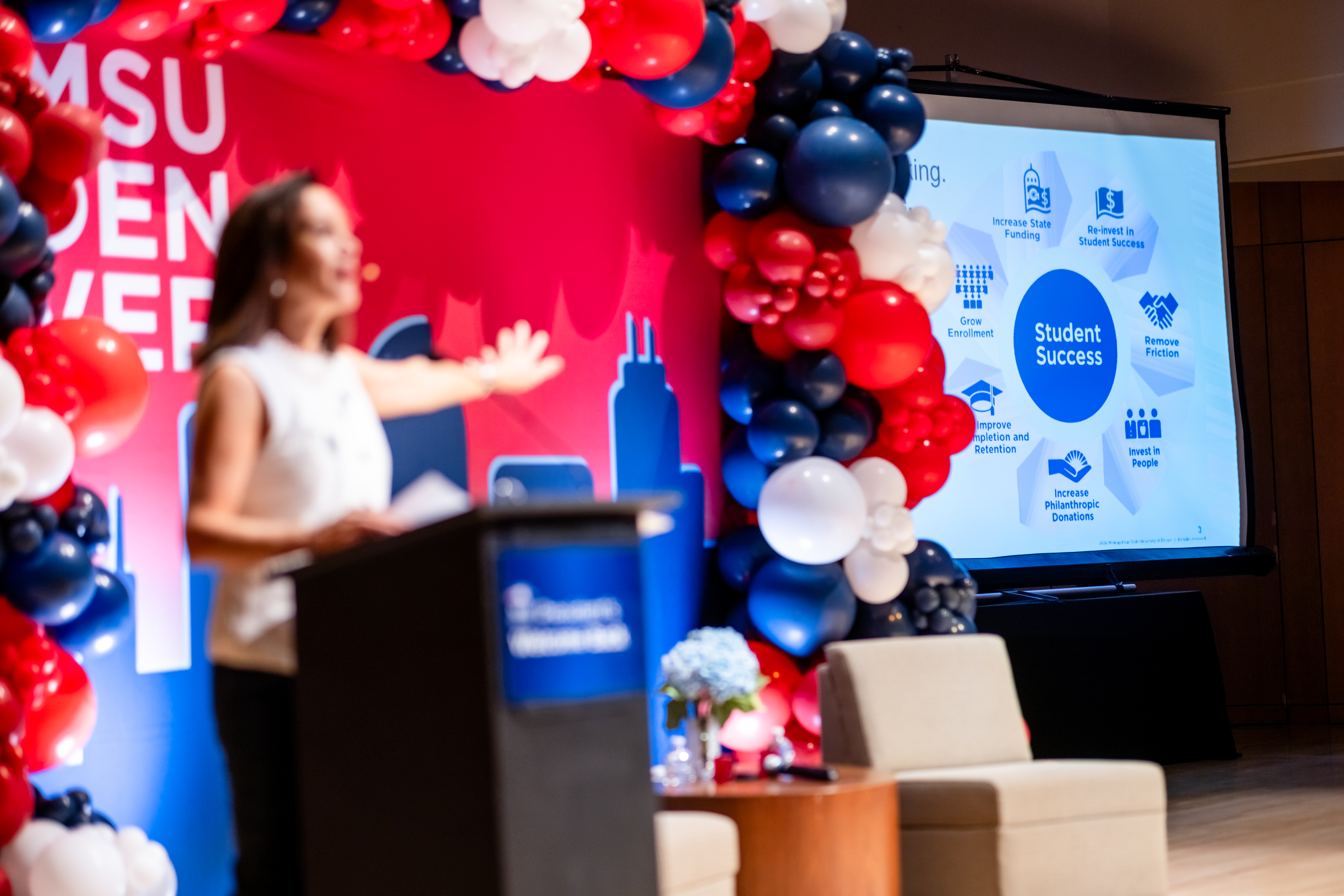 President Janine Davidson speaks on stage while pointing at a screen displaying a slide of the "student-success flywheel," displaying elements representing different faculty and staff roles and functions around a circle.