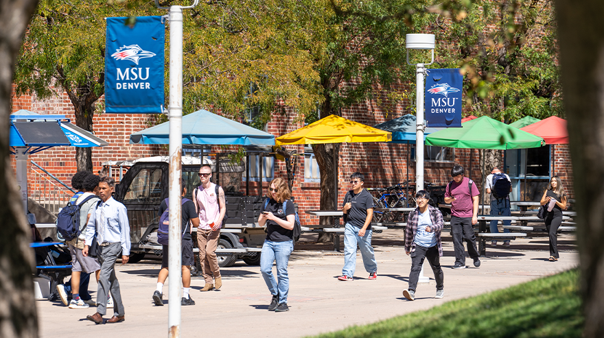 People walk on a campus sidewalk between MSU Denver flags and trees with green and yellow leaves