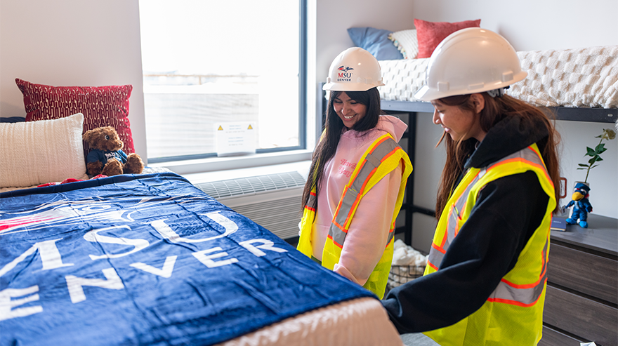 Two students are shown wearing hard hats and reflective vests in a room with two beds and MSU Denver decorations