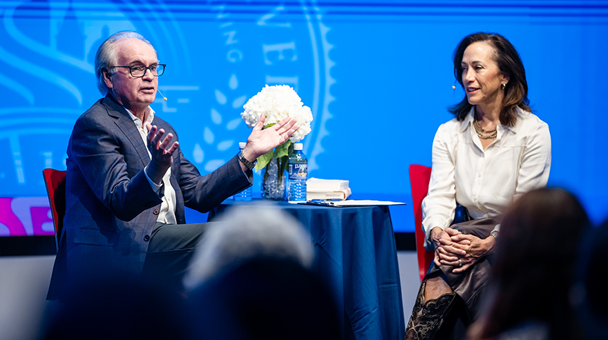 George Sparks and Janine Davidson address a crowd next to a table with flowers and in front of a banner displaying the MSU Denver crest