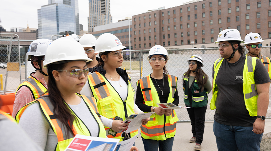 Nine people including student interns and employees stand on a construction site wearing white hard hats, as campus buildings and tall downtown buildings appear in the background