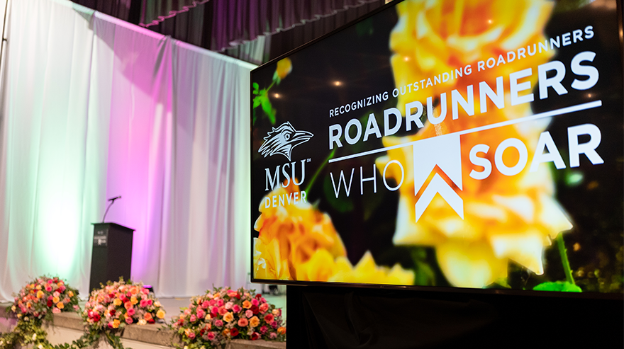 A screen display in front of a stage decorated with flowers shows the logo for Roadrunners Who Soar and moniker "recognizing outstanding Roadrunners"